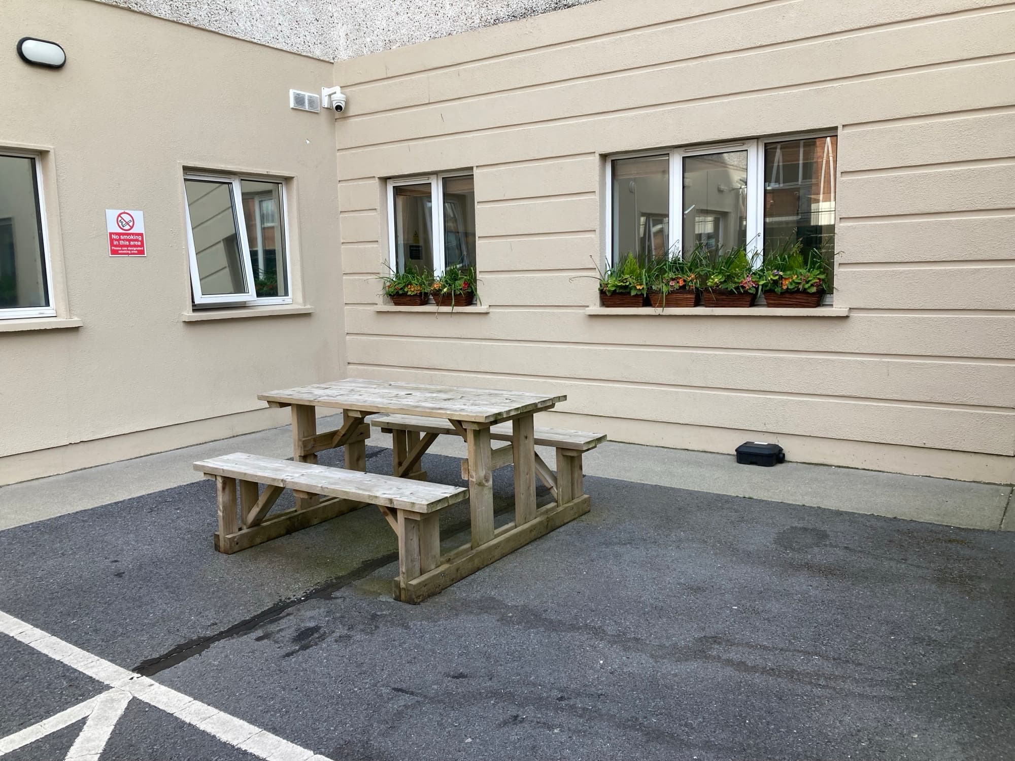 Outdoor seating area with wooden picnic table and benches beside a building courtyard with windows and flower boxes.